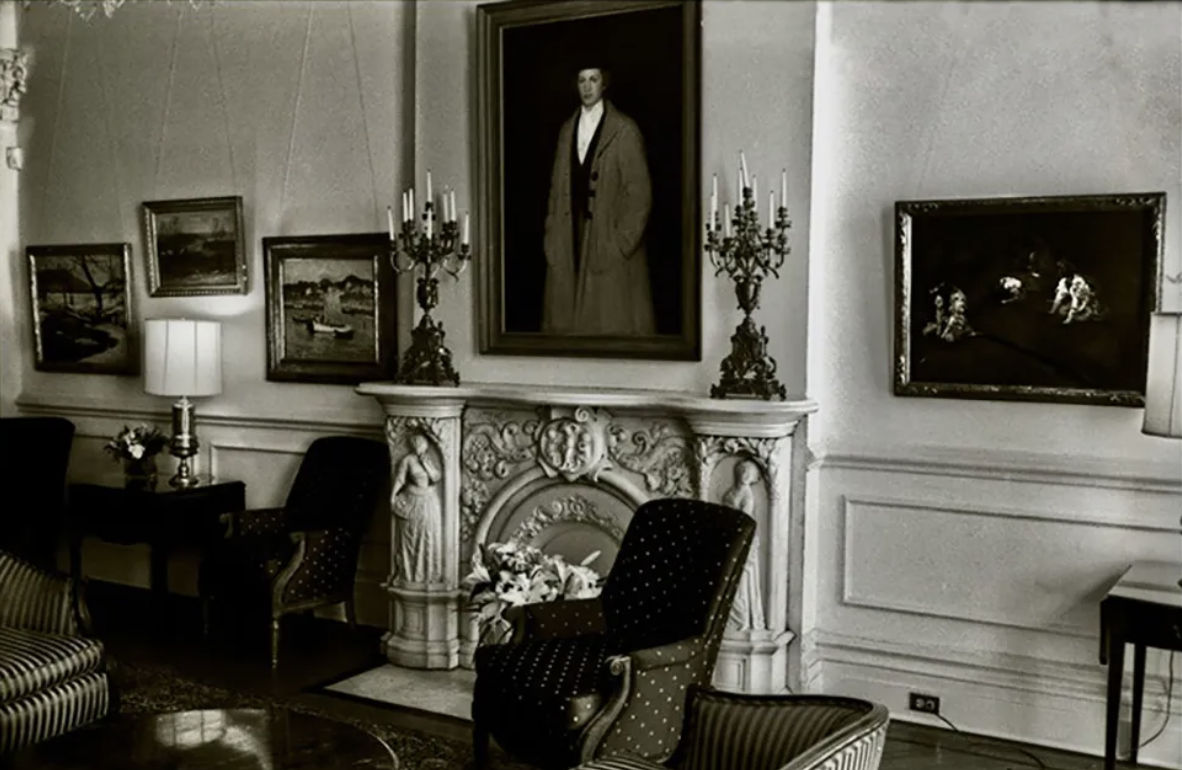 Black and white photograph of old-fashioned living room with fireplace, framed pictures on walls, and furniture