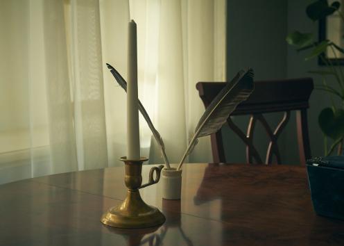 Dark image of a candlestick with candle and quill pen on top of a wooden table with chair, window/curtains, mirror, and plant in backgroun